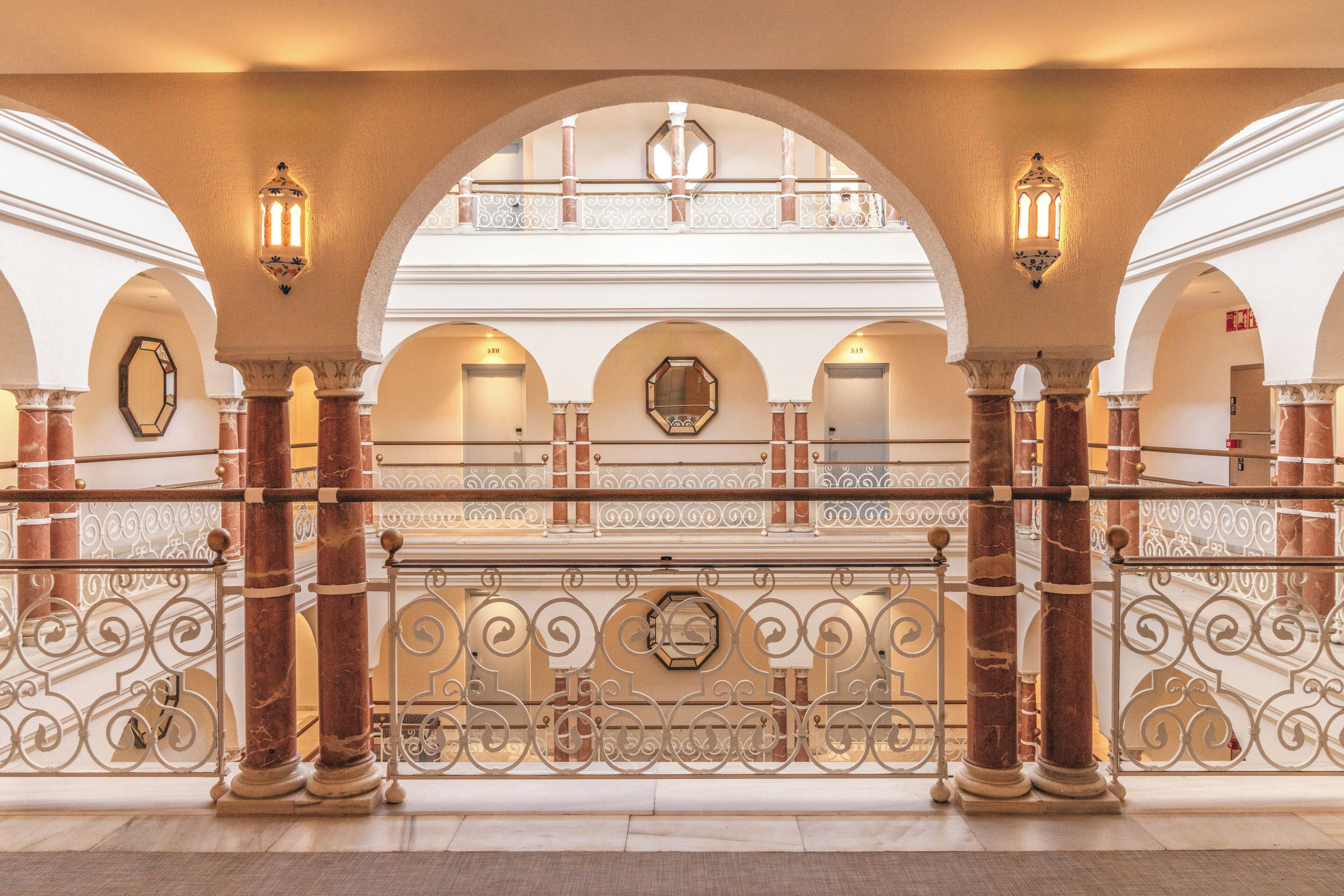 A grand hallway with marble columns, ornate metal railings, and arched doorways.