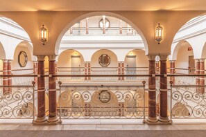 Un gran pasillo con columnas de mármol, barandillas de metal ornamentadas y arcos de entrada.