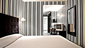 Ein Hotelzimmer mit gestreifter Tapete, einem gepolsterten Kopfteil, einem Bett, einem Schreibtisch mit Lampe und einem Fernseher.