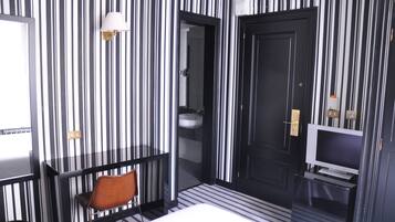 Camera d'albergo con parete a righe bianche e nere, un letto, una TV, un bagno con un lavandino e una sedia.