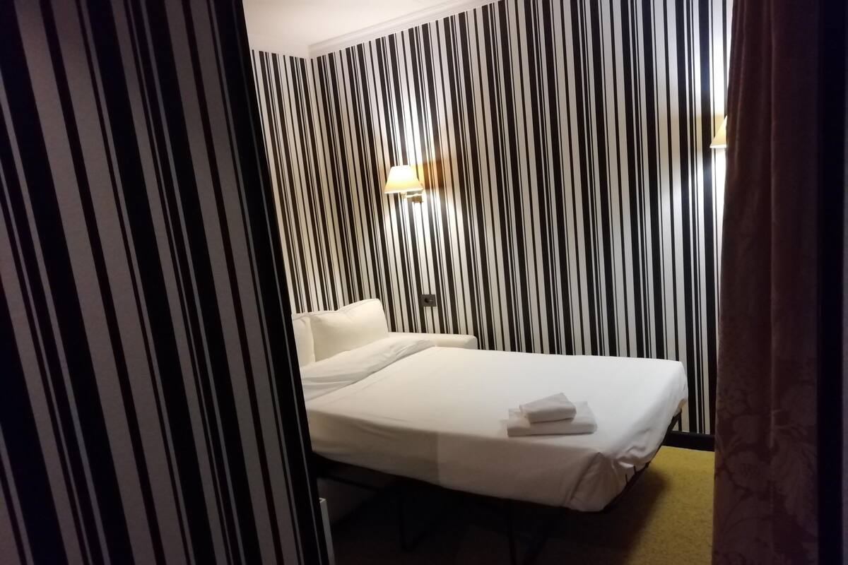Quarto de hotel com cama, mesa de cabeceira, abajur e cortina listrada.