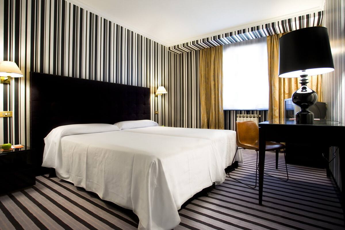 Quarto de hotel com uma cama grande, uma escrivaninha com cadeira, uma luminária e uma janela com cortinas.