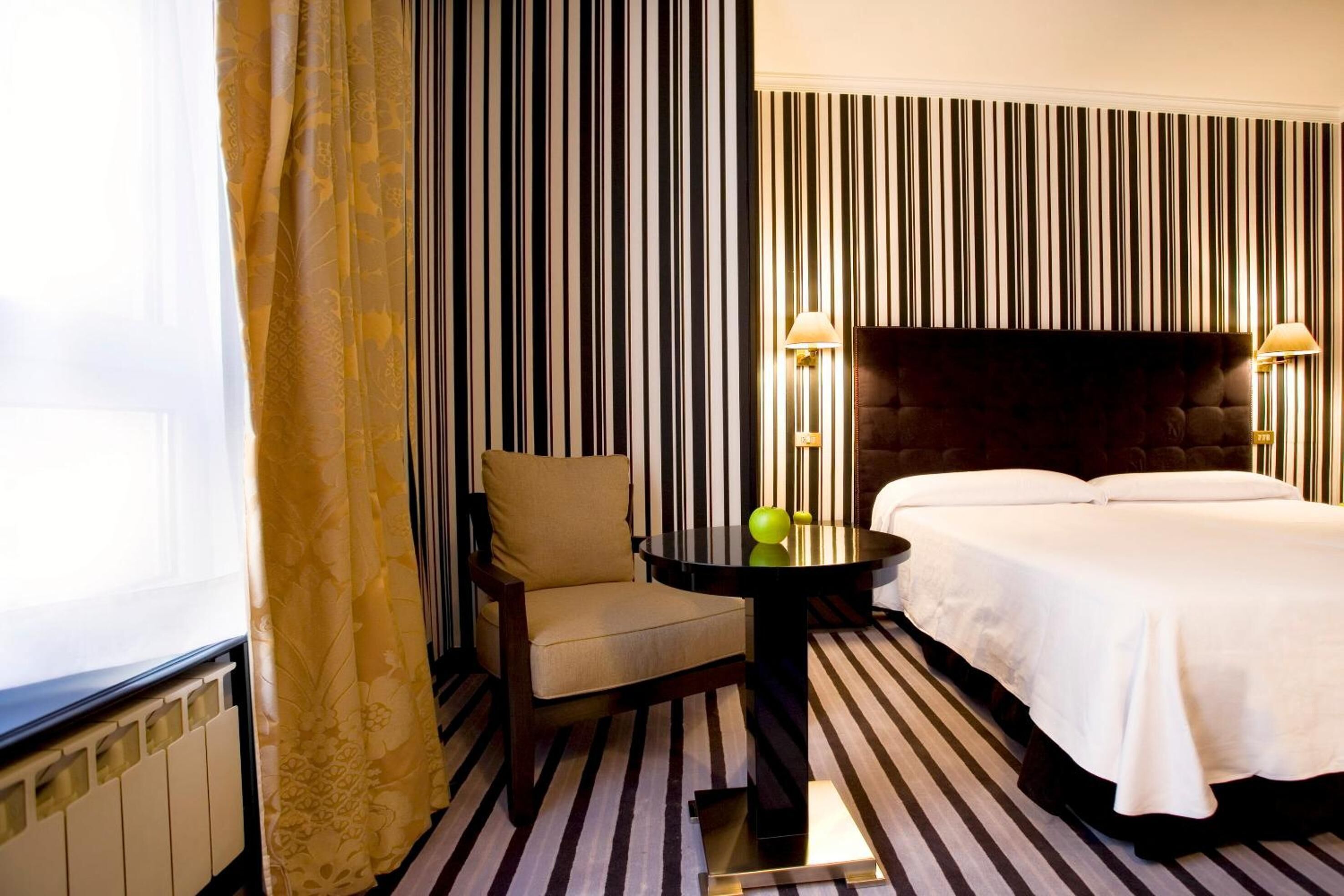 Una habitación de hotel con papel tapiz a rayas, una cama, una silla y una mesa con dos lámparas.
