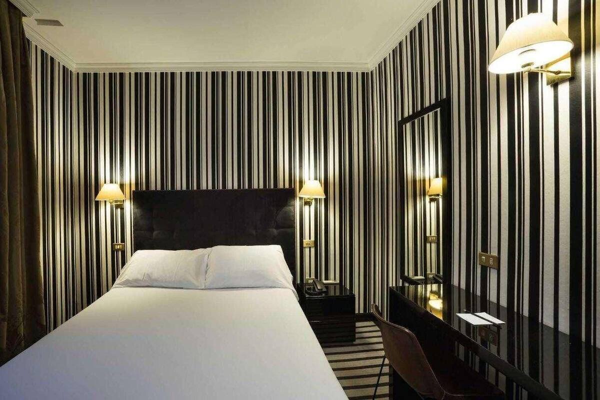 Quarto de hotel com uma cama grande, duas luminárias de cabeceira, uma mesa de apoio, uma cadeira e um espelho na parede.