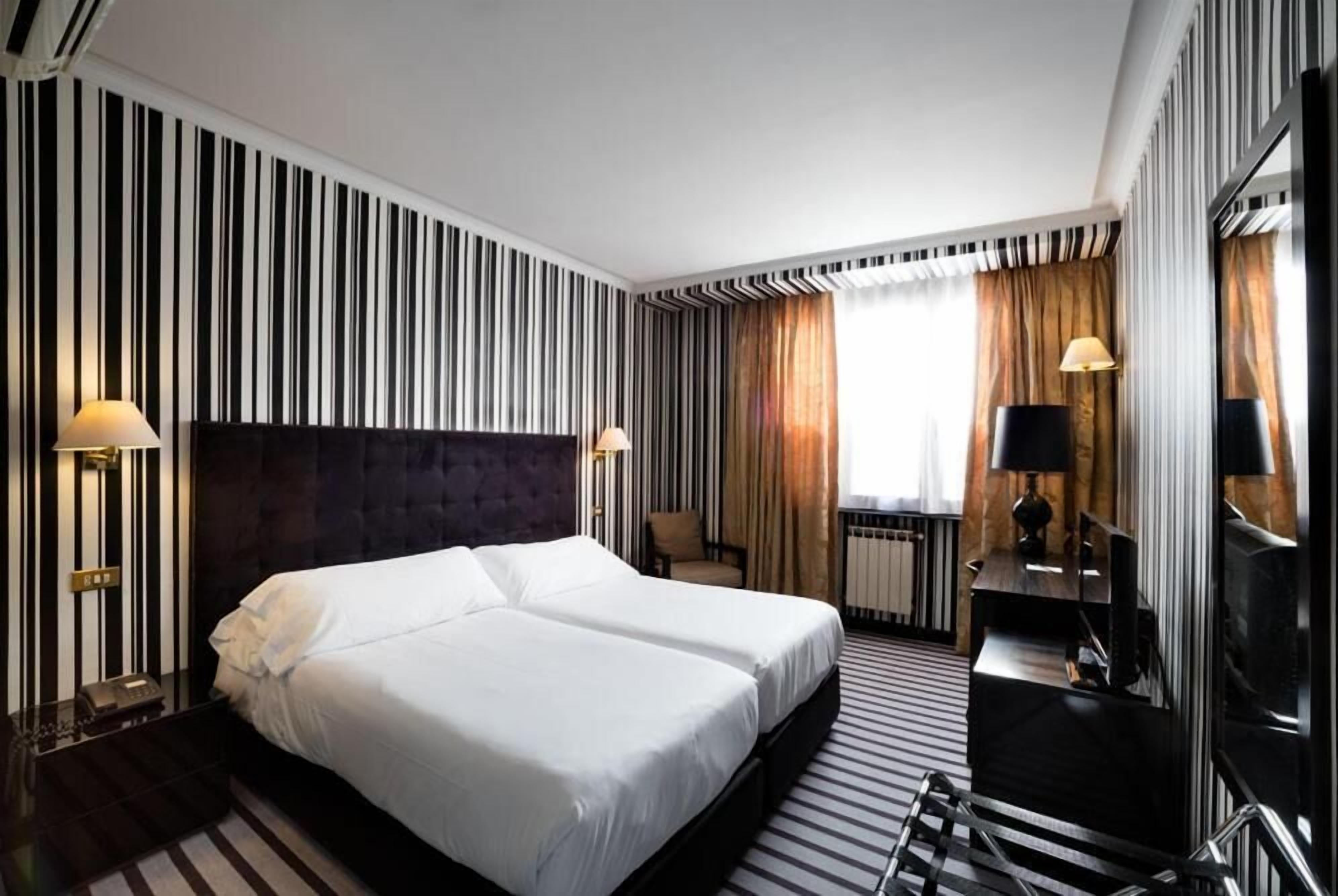 Ein Hotelzimmer mit einem großen Bett, zwei Nachttischen, einem Fernseher und einem Fenster mit Vorhängen.