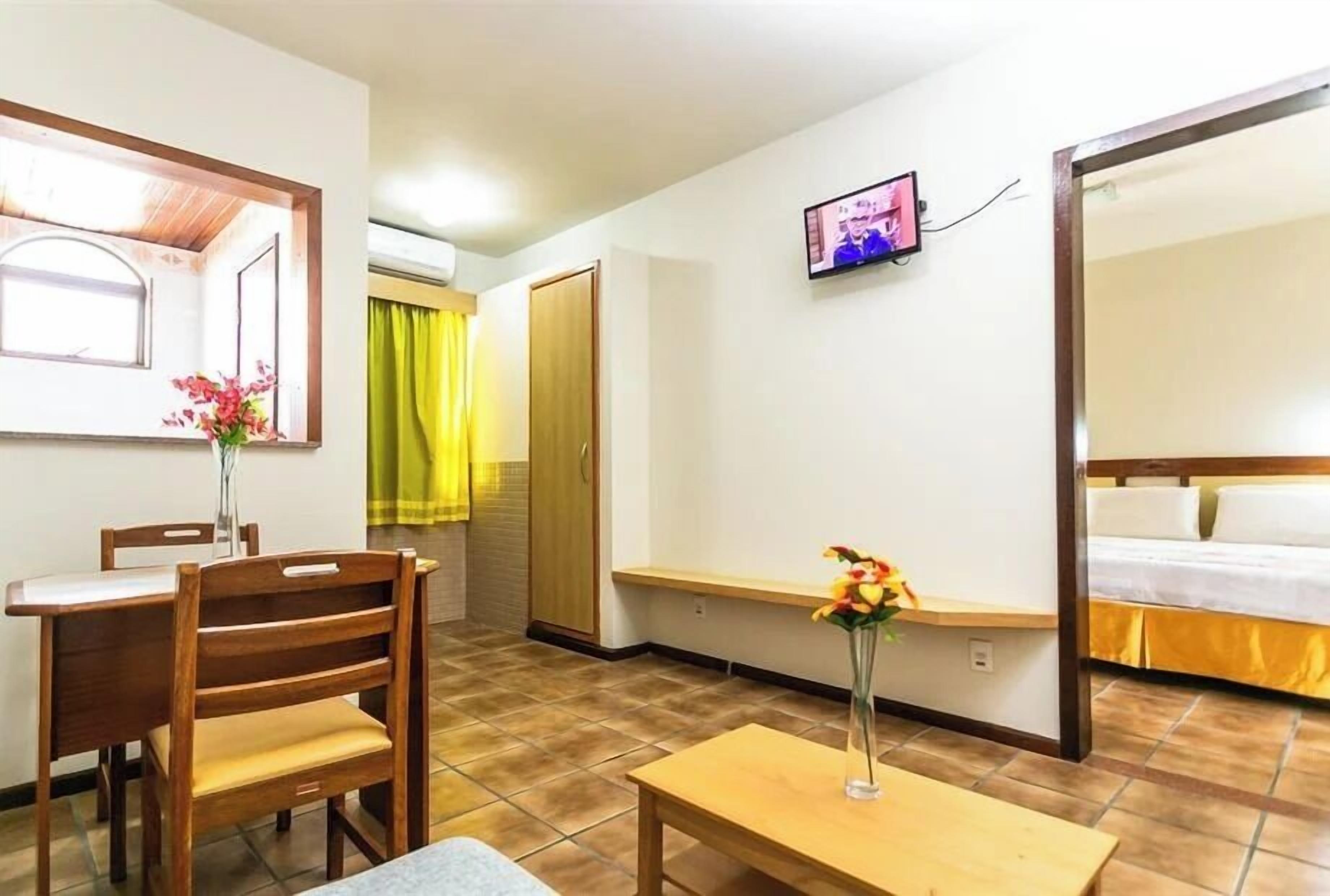 A hotel room with a bed, a TV, a desk with a chair, a vase with flowers, and a window with curtains.