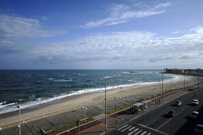 Una carretera costera con playa y océano a un lado, y una acera peatonal y coches aparcados al otro.