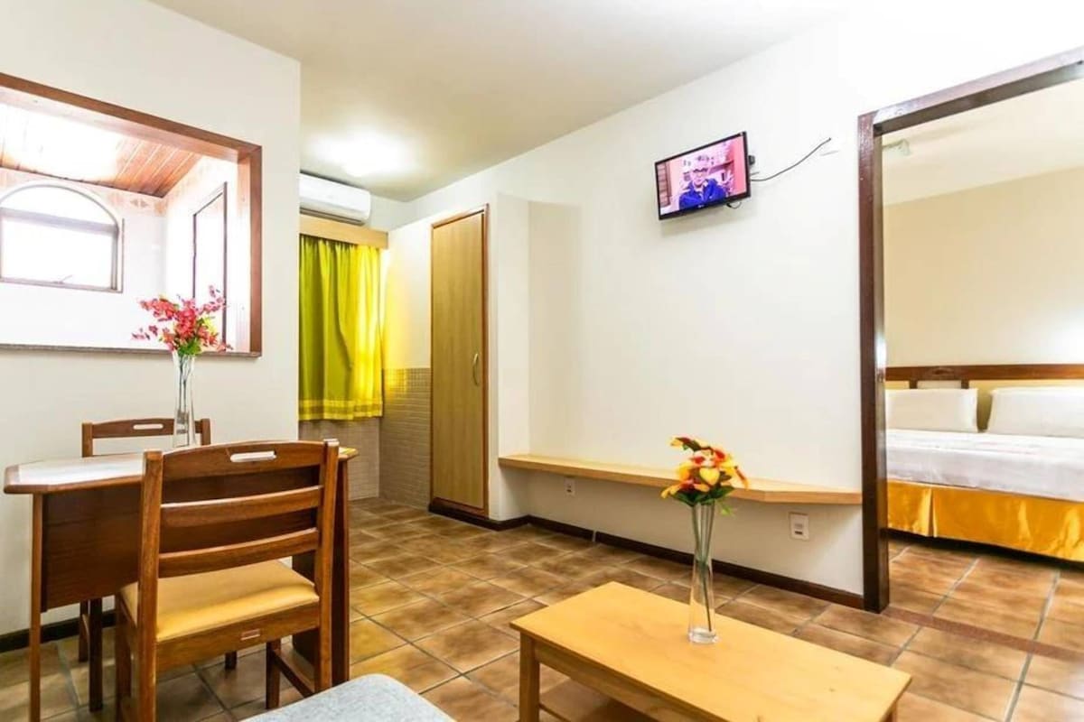 Una habitación de hotel con una cama, un televisor montado en la pared, una mesa de comedor con sillas y una mesita con un jarrón de flores.