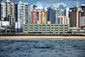 Un paisaje urbano costero con un edificio de varios pisos destacado, con fachada verde y ventanas en arco, junto a una playa de arena y el océano.
