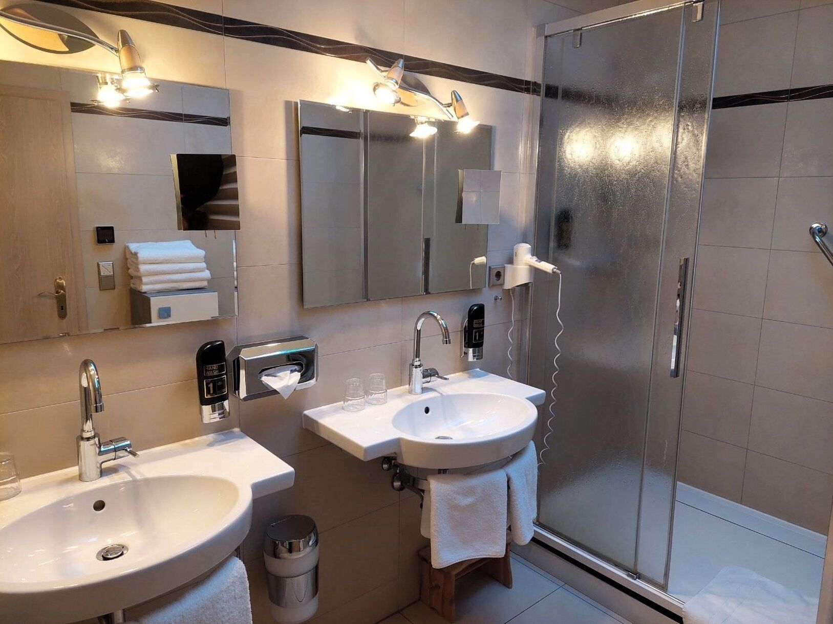Un baño moderno con dos lavamanos, un espejo, un perchero para toallas y una mampara de vidrio para la ducha.
