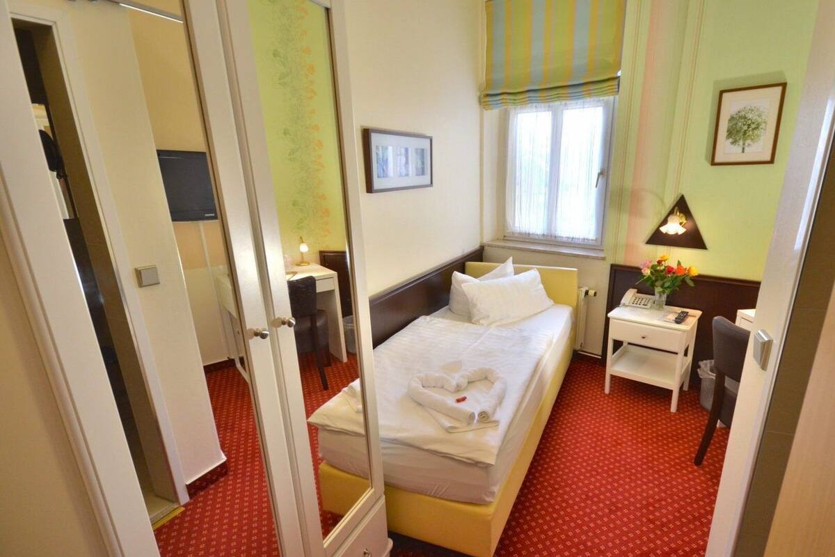A hotel room with a bed, a desk, a chair, a bedside table with a lamp, a window with curtains, and a framed picture on the wall.