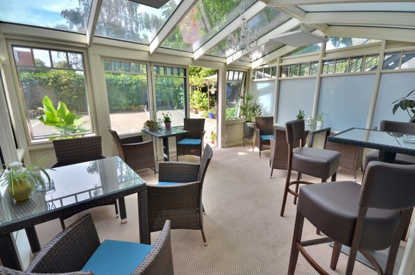 A sunlit patio area with glass walls, wicker furniture, and potted plants.