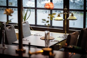A dining area with a table set for two, chairs, a vase with flowers, and a window with a view of trees. - Hotel Alt-Connewitz (Leipzig)