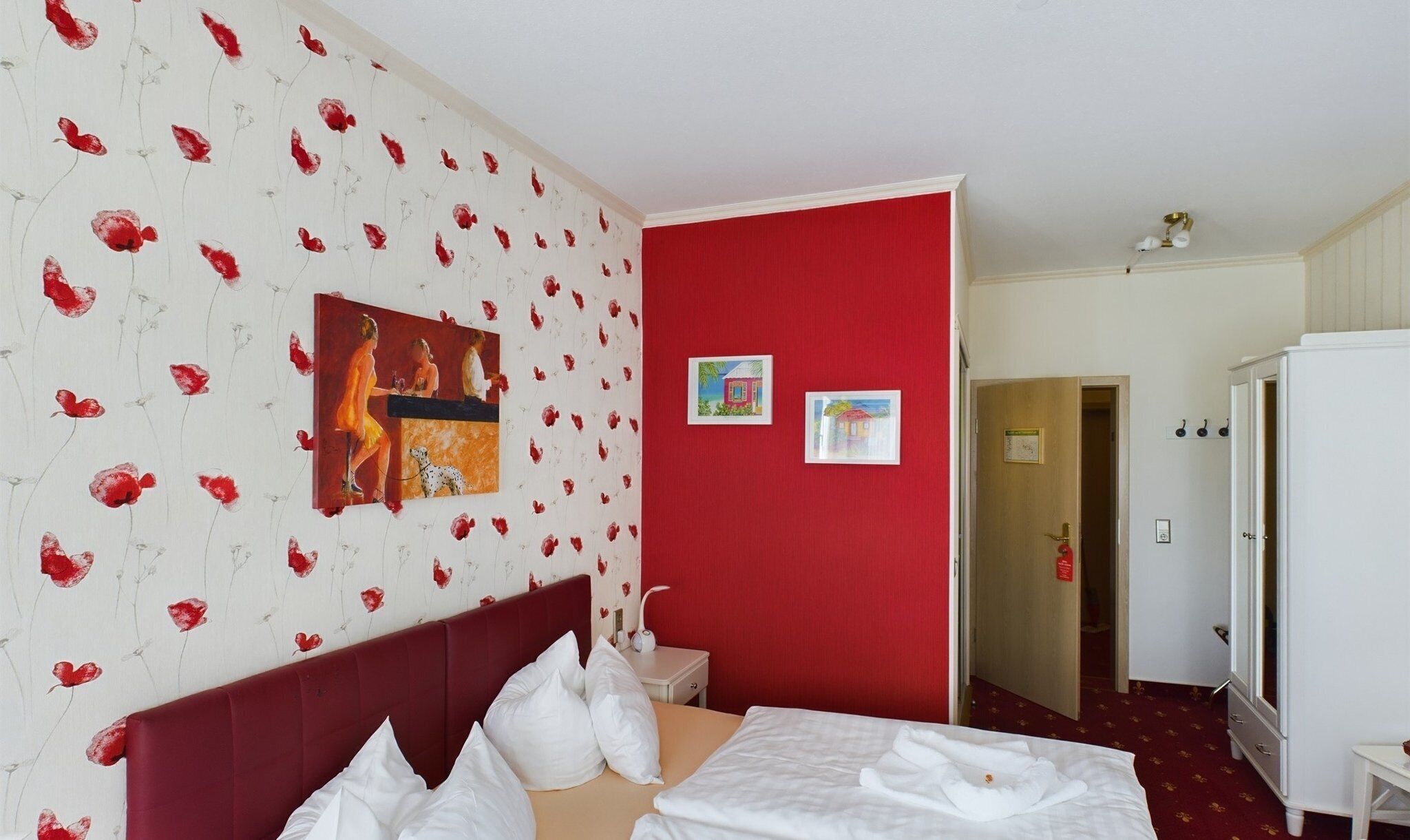 A hotel room with a red accent wall, a bed with white linens, a small bedside table, and a white wardrobe.