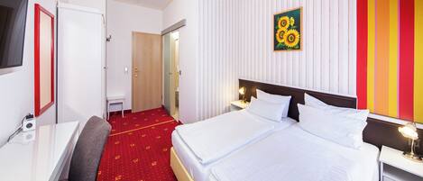 Habitación de hotel con cama, mesita de noche, lámpara y suelo cubierto de alfombra roja.
