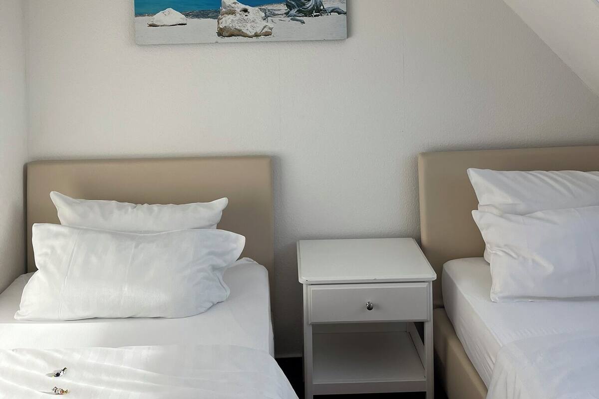 A hotel room with two beds, a nightstand, a painting of a tree by the sea, and a sloped ceiling.