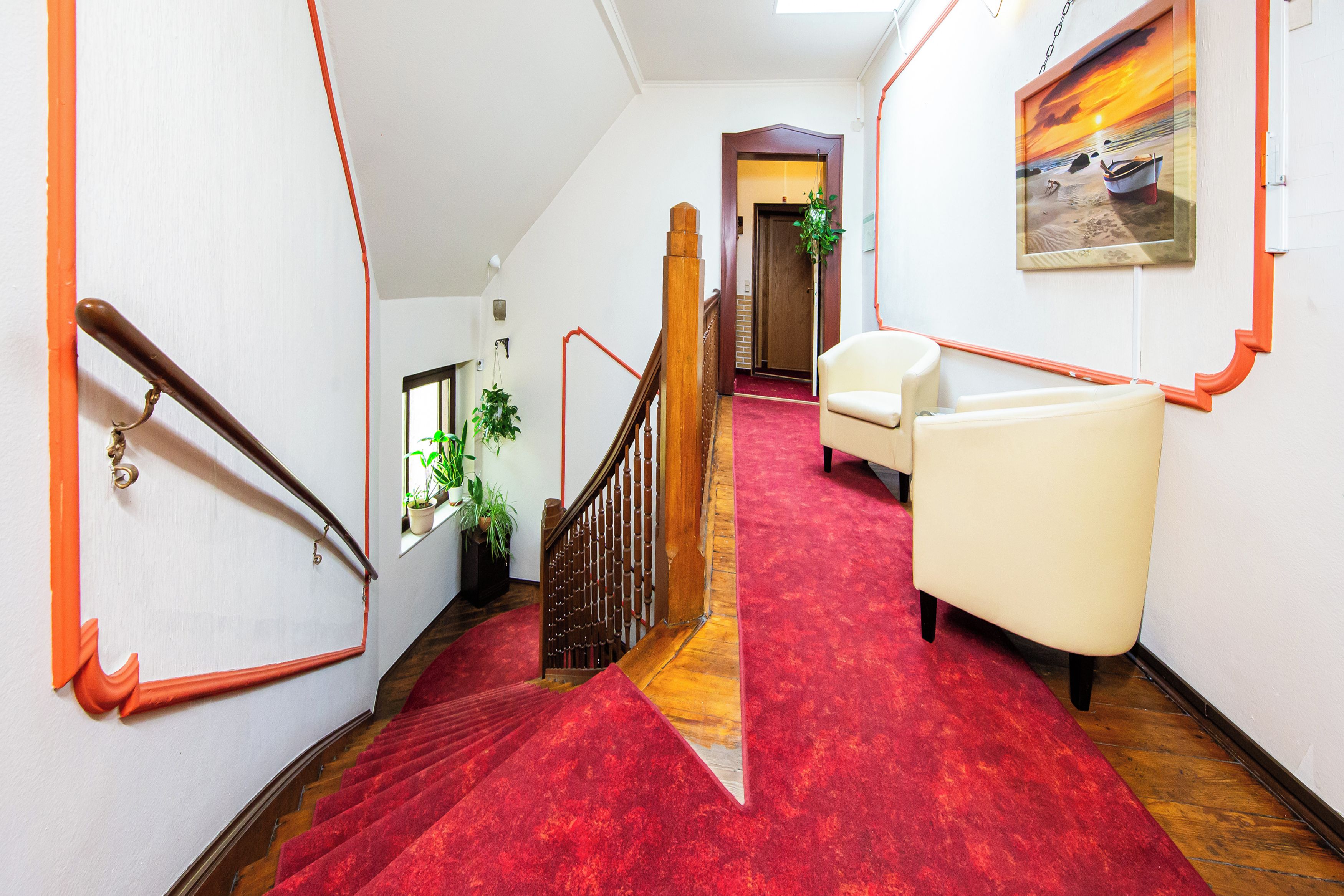 Un pasillo de hotel con alfombra roja, una escalera de madera, un sillón blanco y un cuadro en la pared.