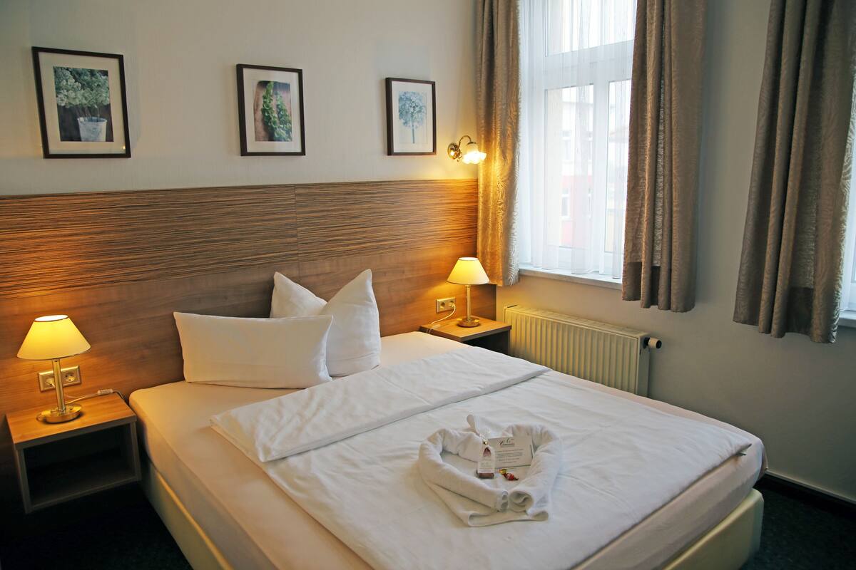 A hotel room with a neatly made bed, two bedside lamps, a framed picture on the wall, and a window with curtains.