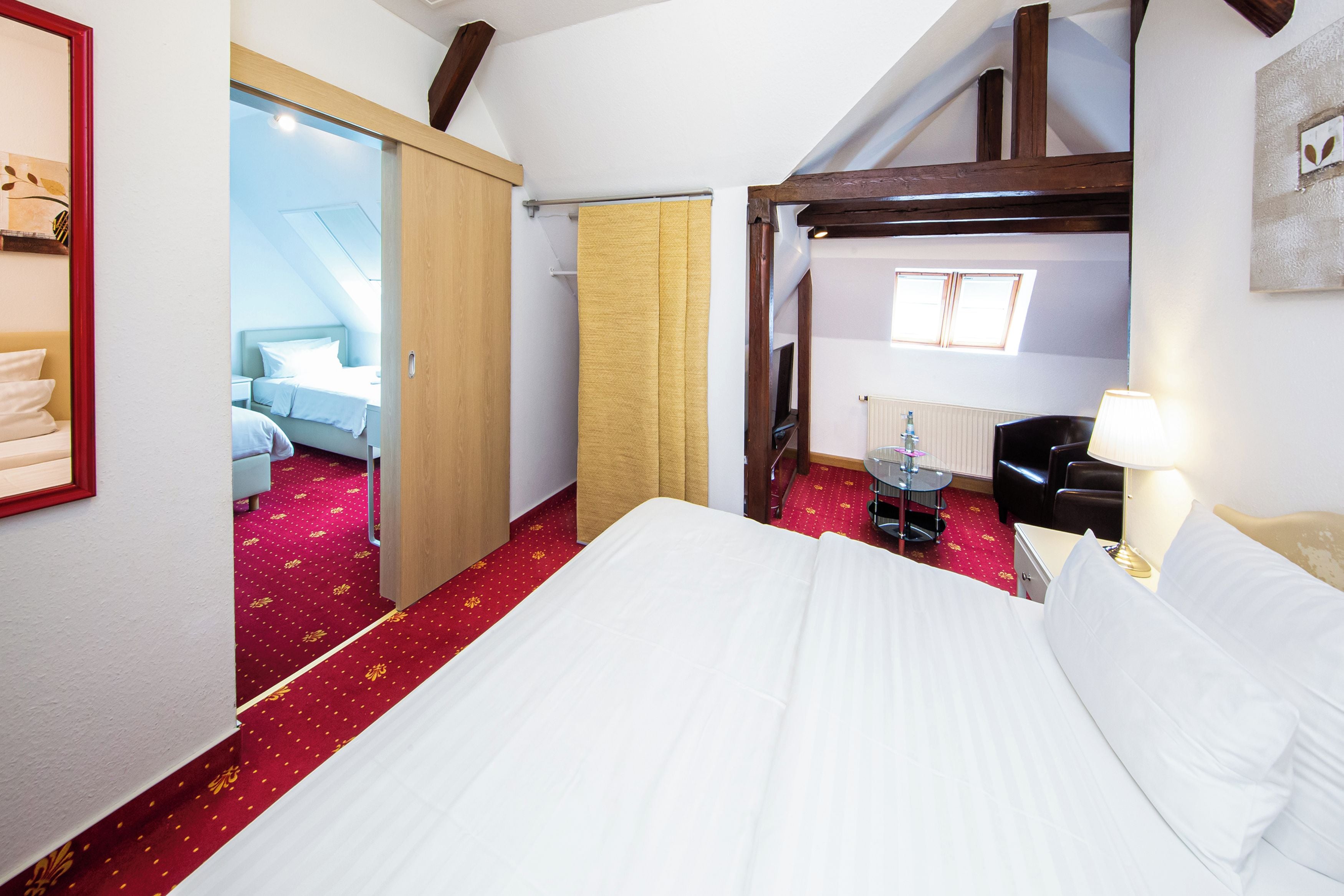 Une chambre d’hôtel avec un grand lit, un bureau avec une lampe, une chaise, une armoire et une vue sur une autre chambre.