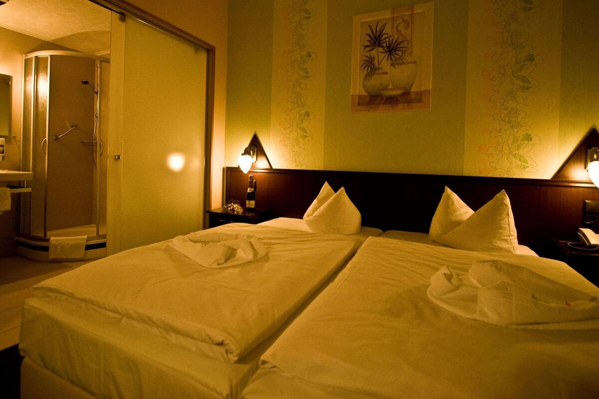 A hotel room with a double bed, a shower, a bedside table with a lamp, and a wall with floral wallpaper.