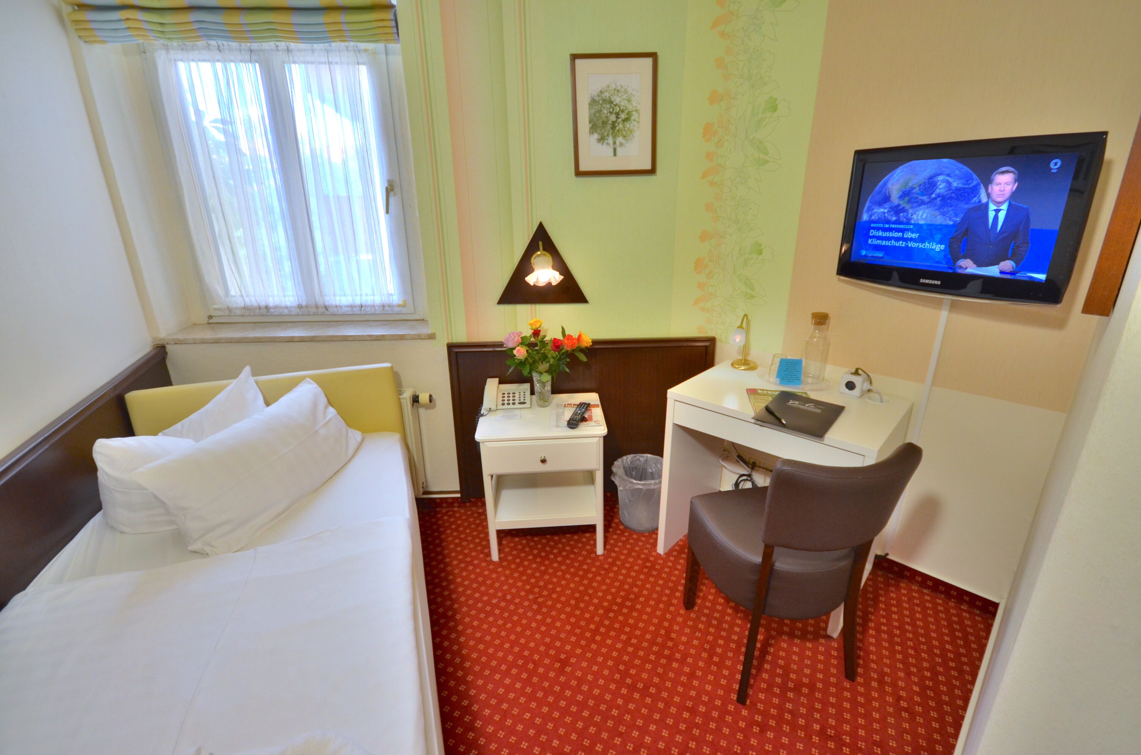 A hotel room with a bed, a desk with a chair, a TV, a lamp, and a small trash bin.