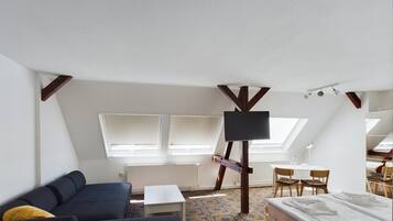 Habitación pequeña y bien iluminada con sofá, cama, una mesita y un televisor montado sobre un soporte de madera.