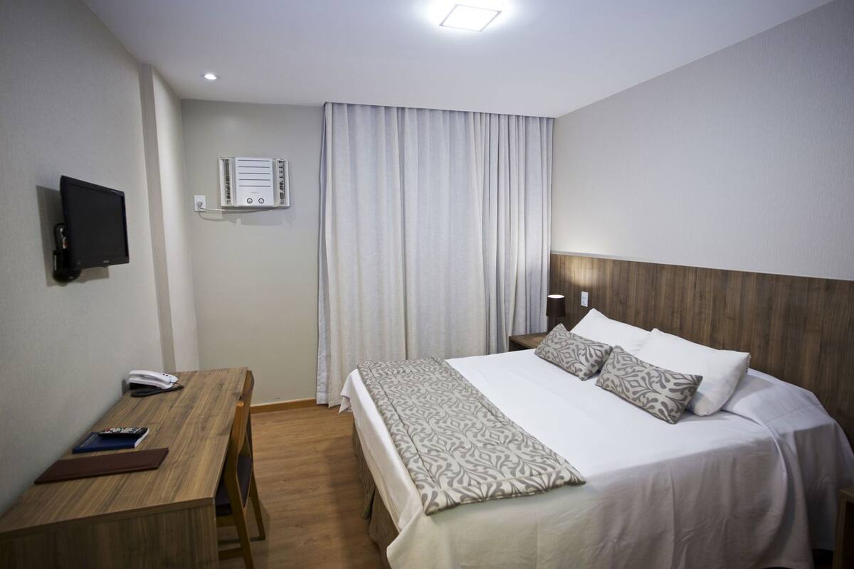 A hotel room with a bed, bedside table, desk, chair, TV, air conditioning unit, and curtains.