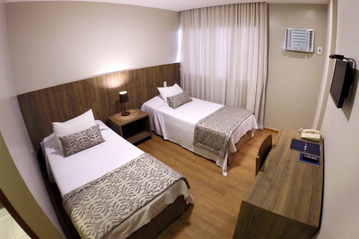 A hotel room with two beds, a desk, a lamp, a telephone, and a wall-mounted air conditioner.