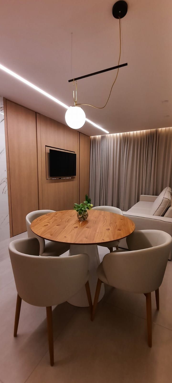 A modern living room with a round wooden dining table, beige chairs, a flat-screen TV mounted on the wall, and a ceiling light fixture.