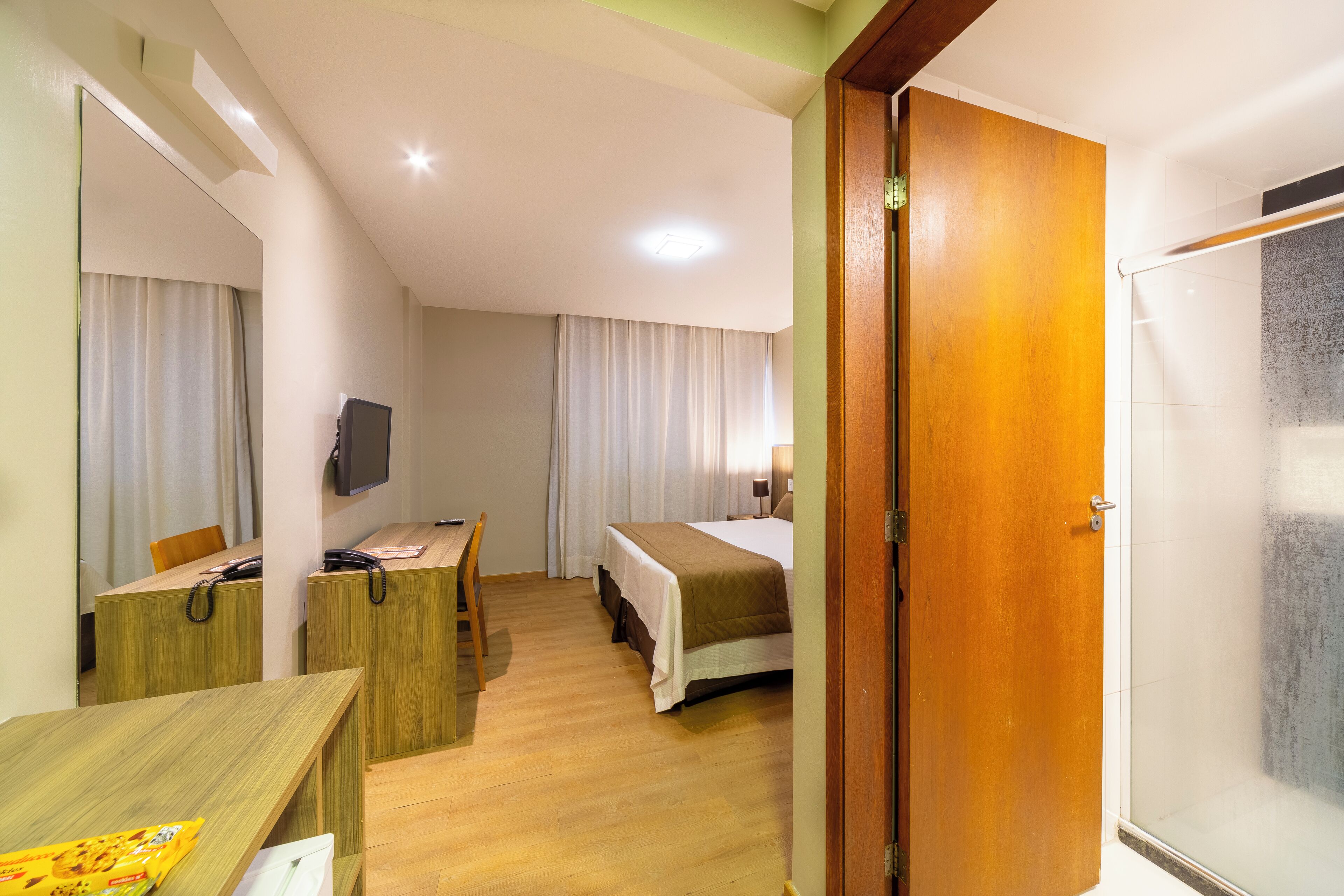 Una habitación de hotel con un escritorio de madera, una cama con colcha blanca y marrón, un televisor en la pared y una puerta que da a la zona de la ducha.