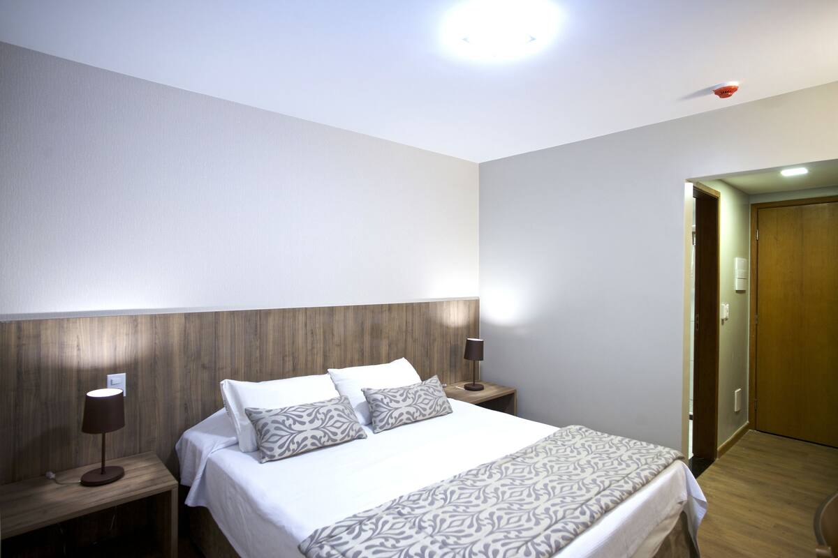 A bedroom with a wooden headboard, a bed with patterned bedding, two bedside lamps, and a door leading to another room.