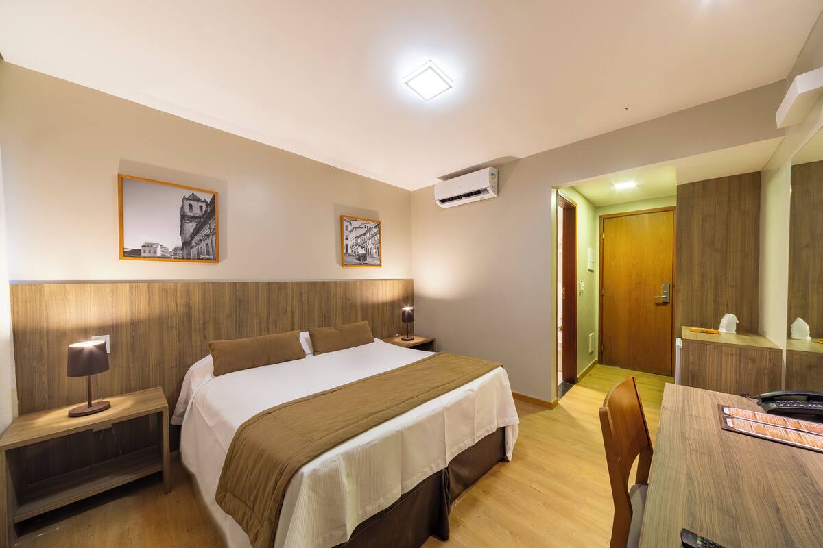 A hotel room with a bed, bedside tables, a desk, and a chair. There is a door leading to another room.