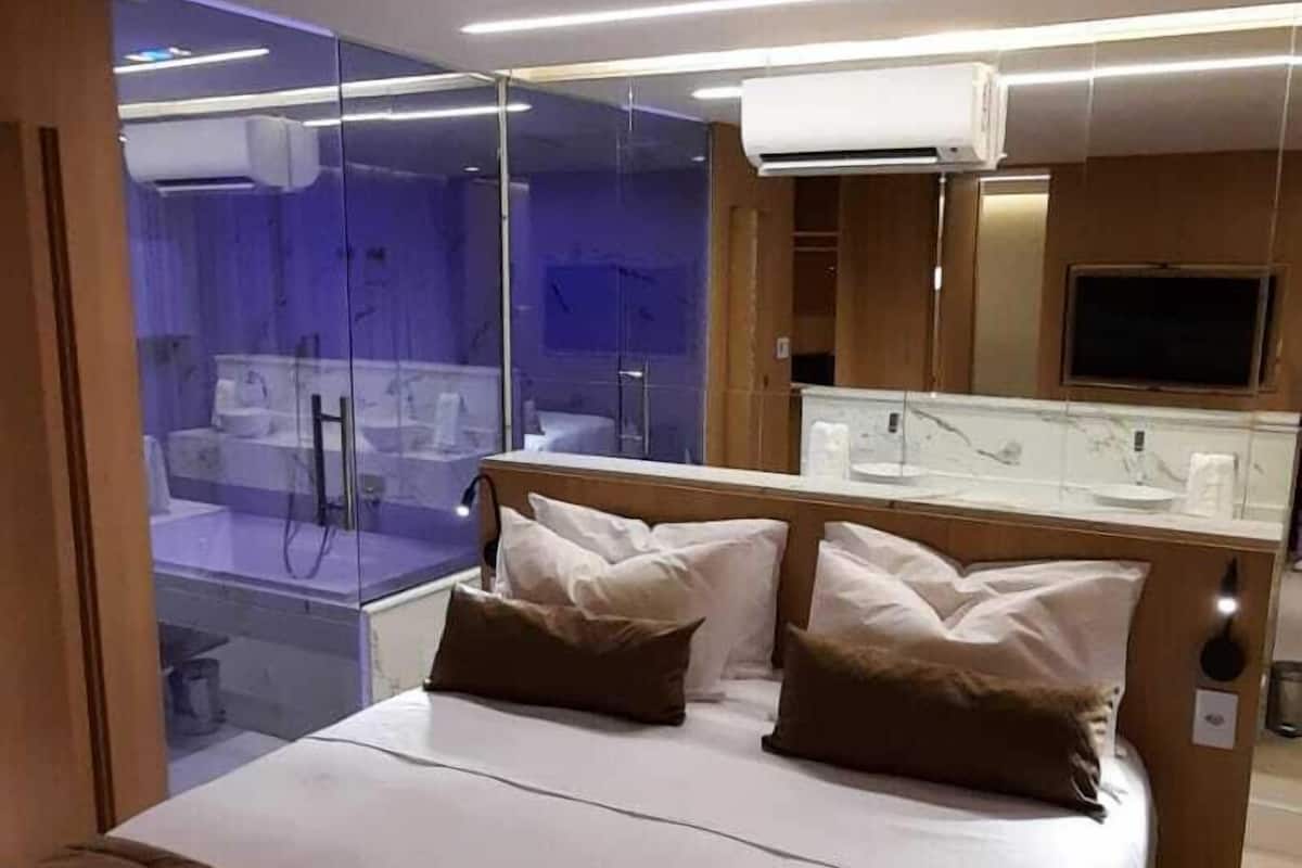 A modern hotel room with a large bed, a glass-enclosed shower area, a television, and a minibar.