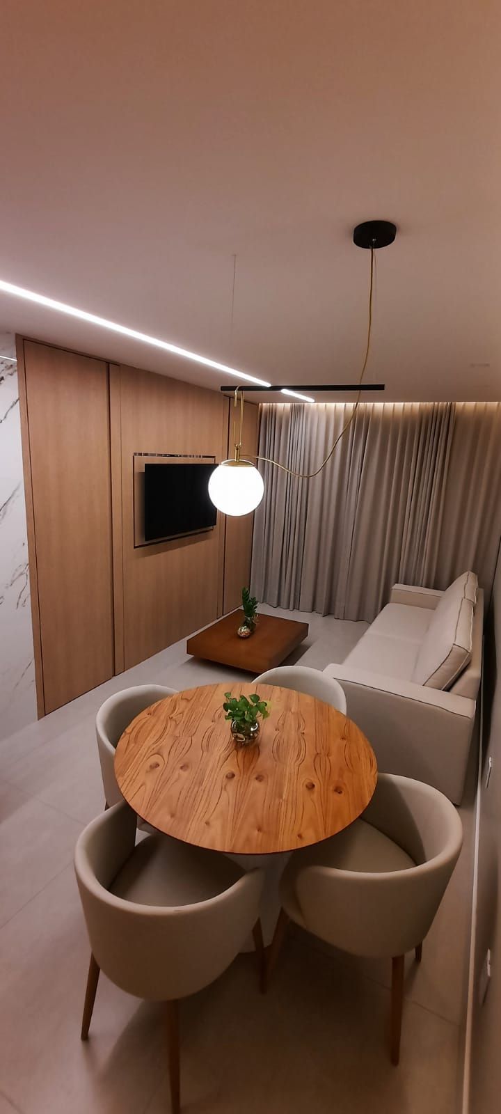 A modern room with a round wooden table, two chairs, a flat-screen TV mounted on the wall, a ceiling light fixture, and a sofa with curtains.