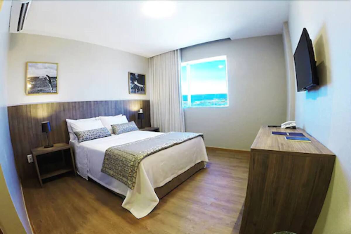 A hotel room with a bed, bedside tables, a desk, a trash bin, a window with a view, and a mounted TV.