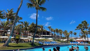 A swimming pool area with lounge chairs and umbrellas, surrounded by palm trees and a building with a red roof. - EL ARAM IMIRÁ BEACH RESORT (Natal)