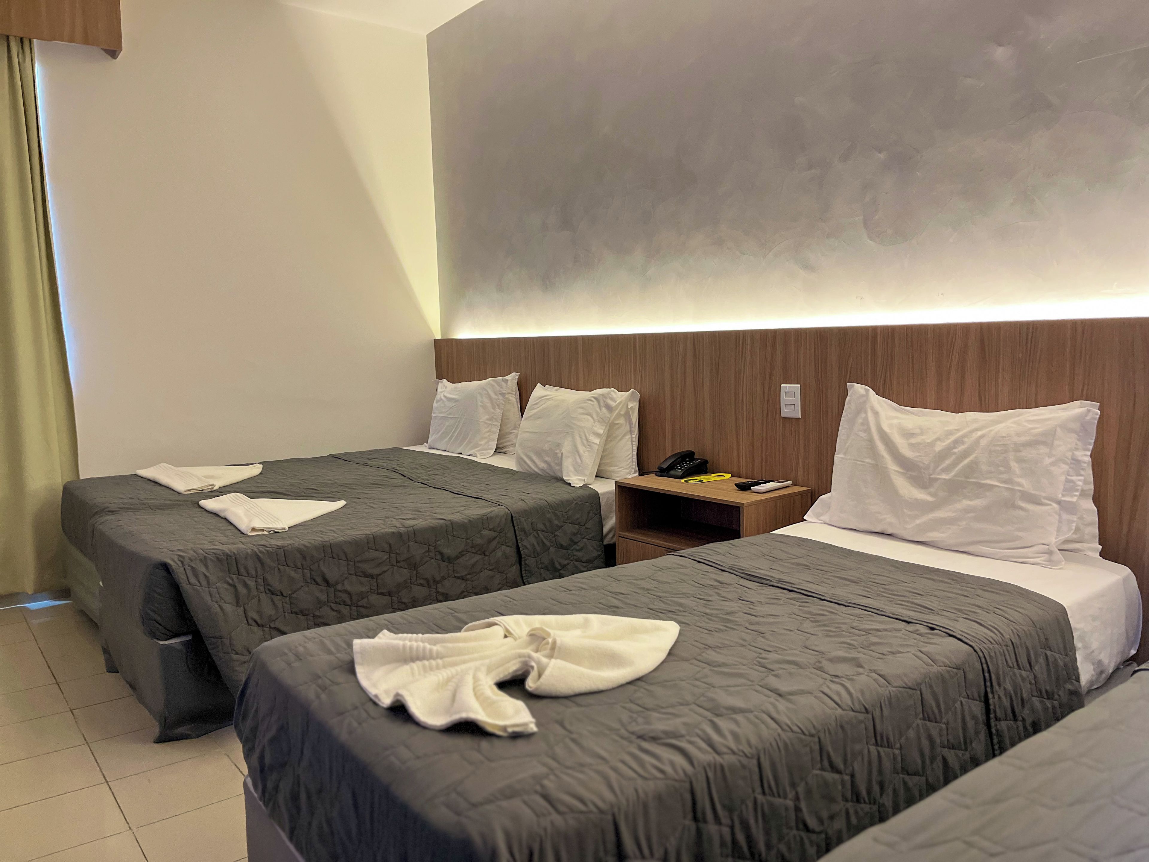 Quarto de hotel com duas camas, mesa de cabeceira com telefone e controle remoto, e luminária fixada na parede.