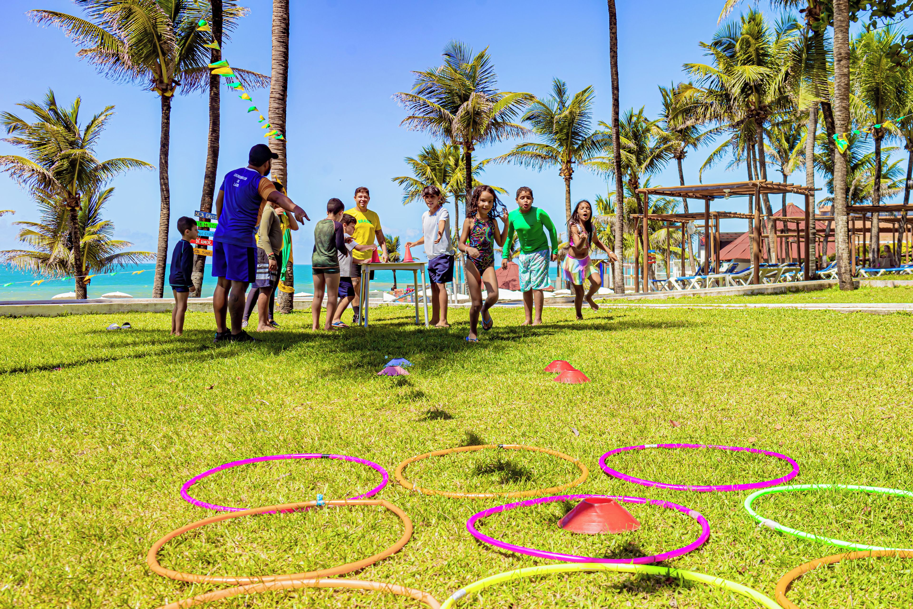 Un grupo de personas jugando un juego en un área verde con aros de colores, palmeras y una playa al fondo.