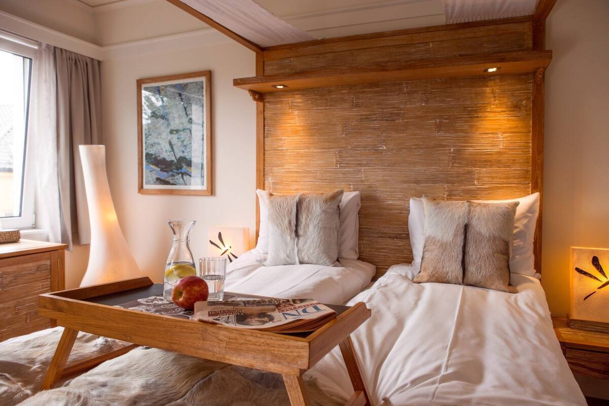 A hotel room with a wooden headboard, two white beds, a wooden side table with a tray, a lamp, and a framed picture on the wall.