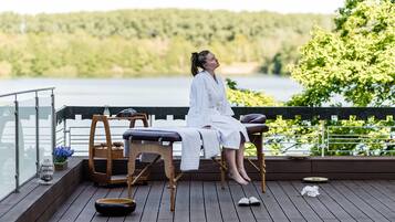 A person seated on a wooden deck with a massage table, overlooking a body of water and greenery.