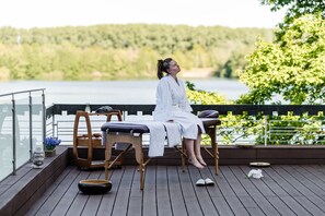 Een persoon zittend op een houten terras met een massagetafel, met uitzicht op water en begroeiing.