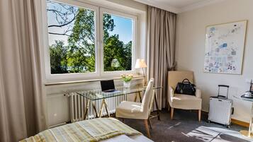 A hotel room with a bed, a desk with a laptop, a chair, a radiator, a window with curtains, and a map on the wall.