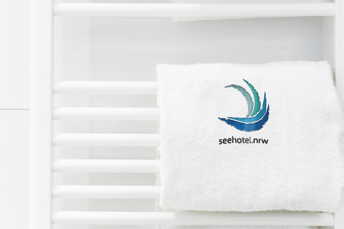 Een witte handdoekhouder met twee witte handdoeken, elk voorzien van een logo en de tekst 'seehotel.nrw'.