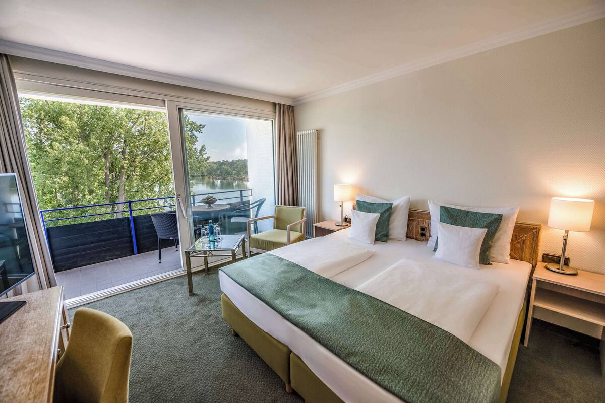 Hotelkamer met een groot bed, twee nachtkastjes met lampen, een bureau met stoel, een balkon met tafel en stoelen, en uitzicht op bomen en een waterpartij.