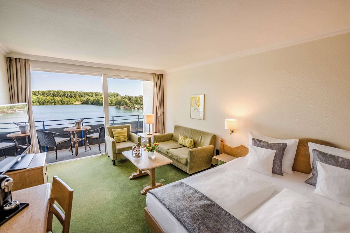 Een hotelkamer met een bed, een bank, een eettafel en een balkon met uitzicht op een meer.
