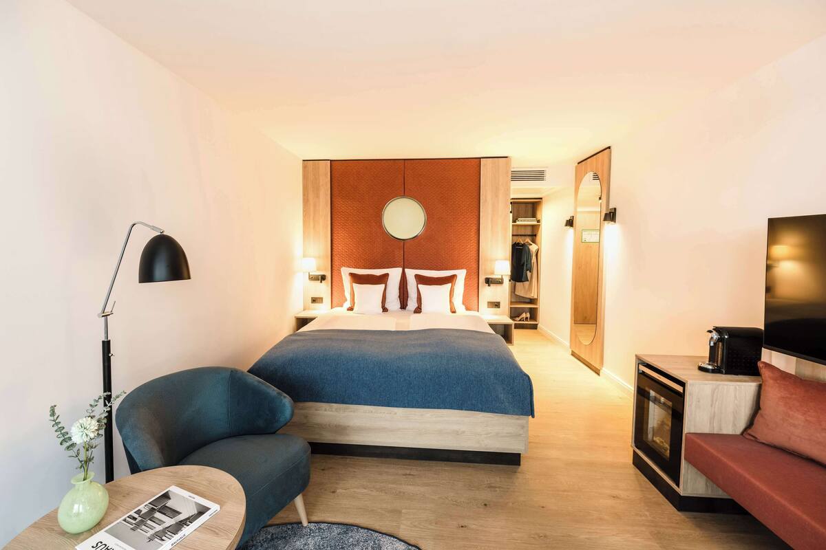 Een moderne hotelkamer met een groot bed, een zithoek met een bank en fauteuil, een klein tafeltje met een vaas en tijdschrift, en een televisie op een standaard.