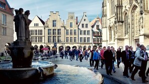 A historic square with a fountain, people walking, and traditional buildings.
