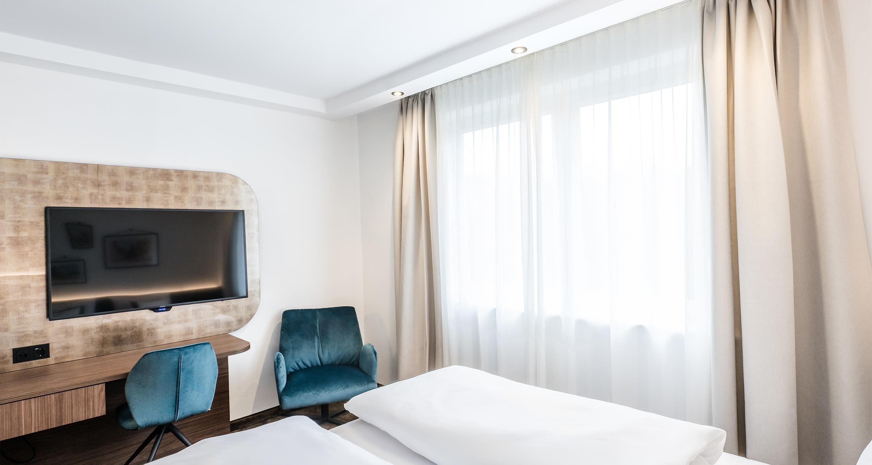 Habitación de hotel con un televisor de pantalla plana montado en un panel de madera, un sillón azul, cama con ropa blanca y un ventanal con cortinas traslúcidas.