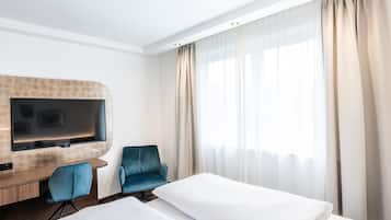 Habitación de hotel con un televisor de pantalla plana montado en un panel de madera, un sillón azul, cama con ropa blanca y un ventanal con cortinas traslúcidas.
