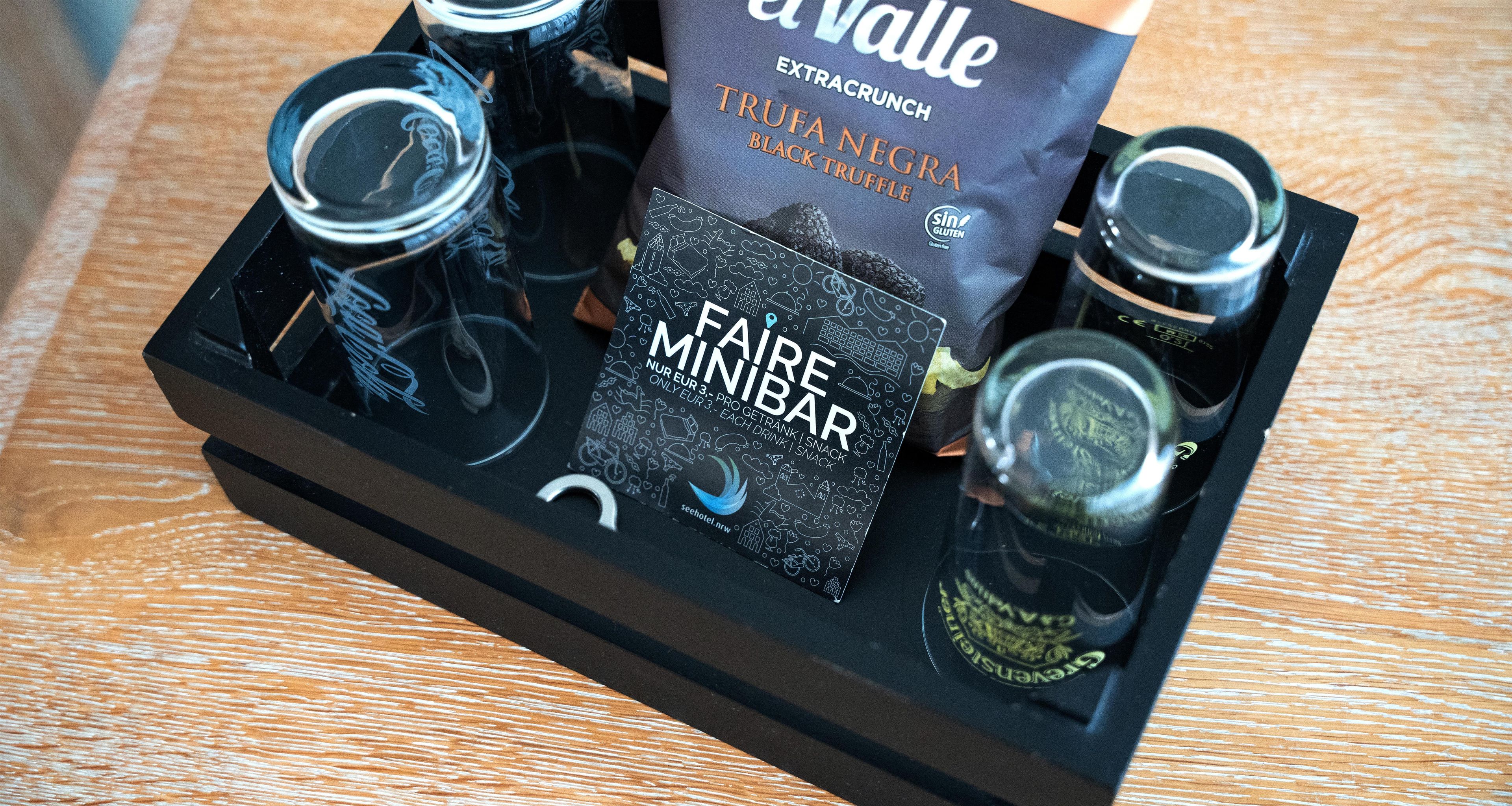 Una bandeja negra con un paquete de chocolate de trufa, una caja con la etiqueta 'FAIR MINIBAR' y dos botellas.